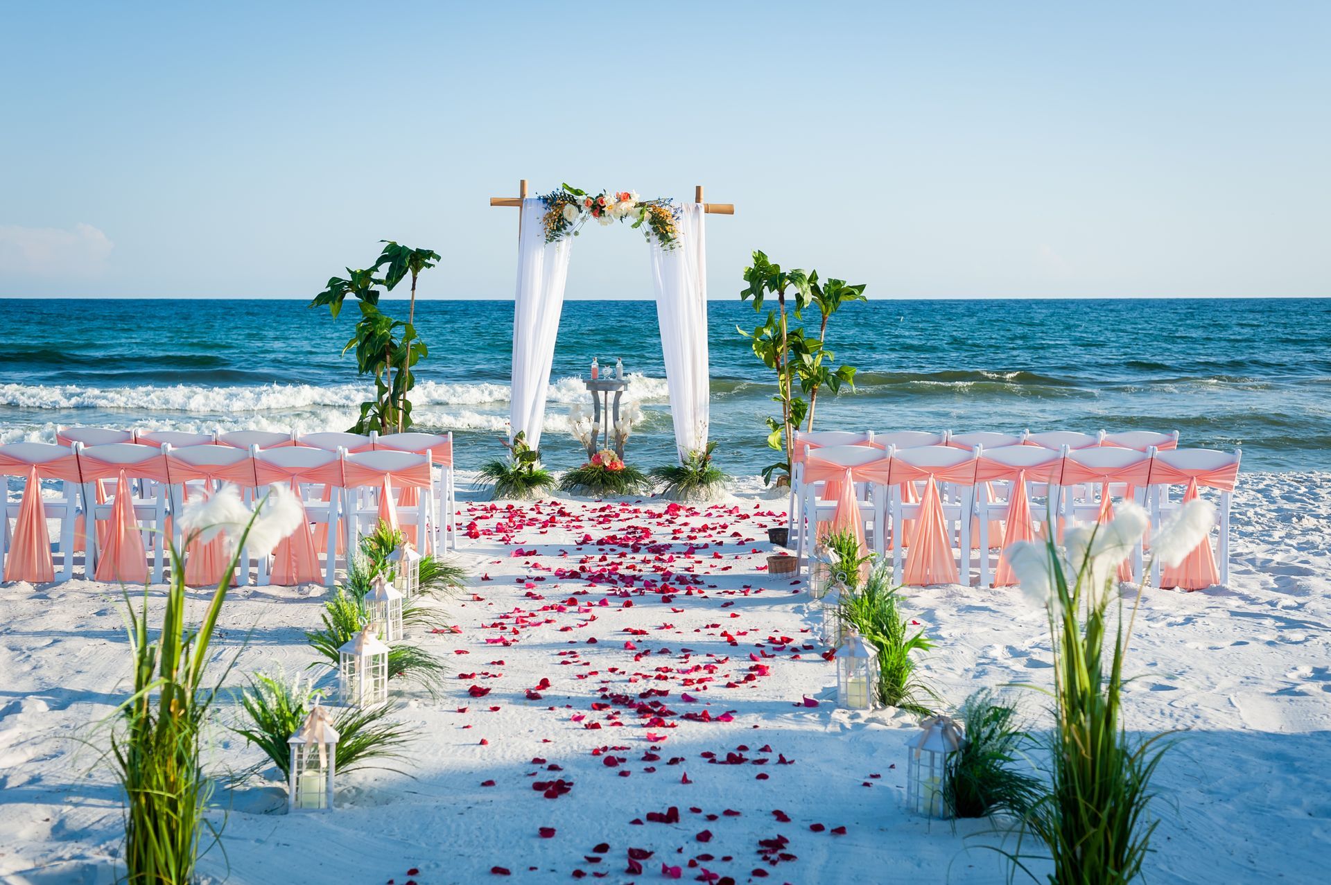 Beach wedding aisle with white drapery, pink chairs, flower petals, and ocean backdrop
