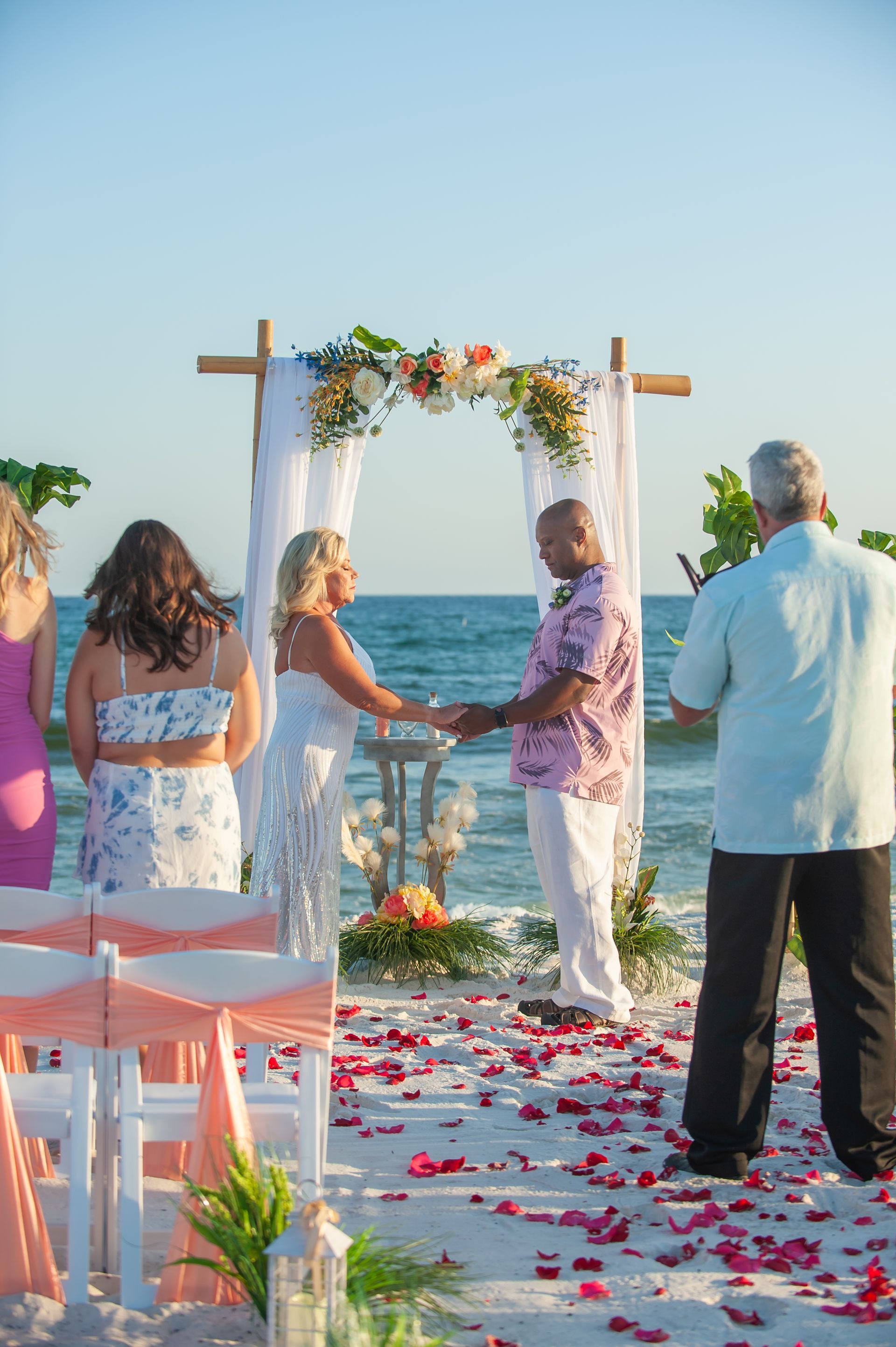 Couple holding hands under a floral arch at a beach wedding ceremony with guests nearby