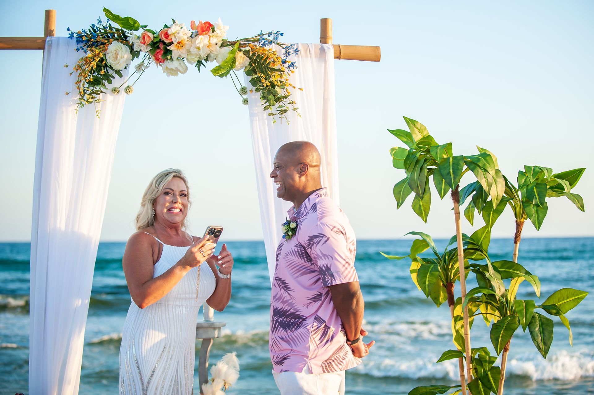 Couple exchanging rings under a flower arch on a beach at sunset