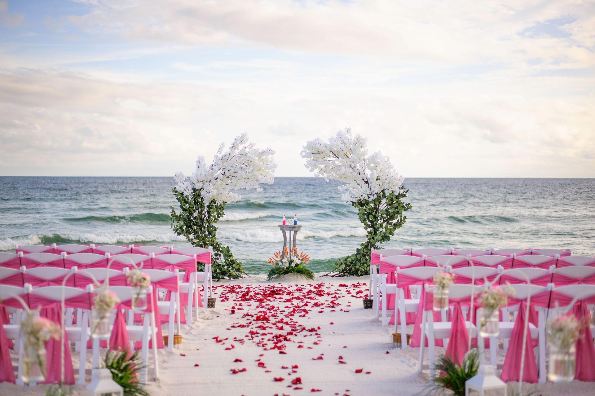 Beach wedding aisle with pink chairs, rose petals, and floral arch by the ocean
