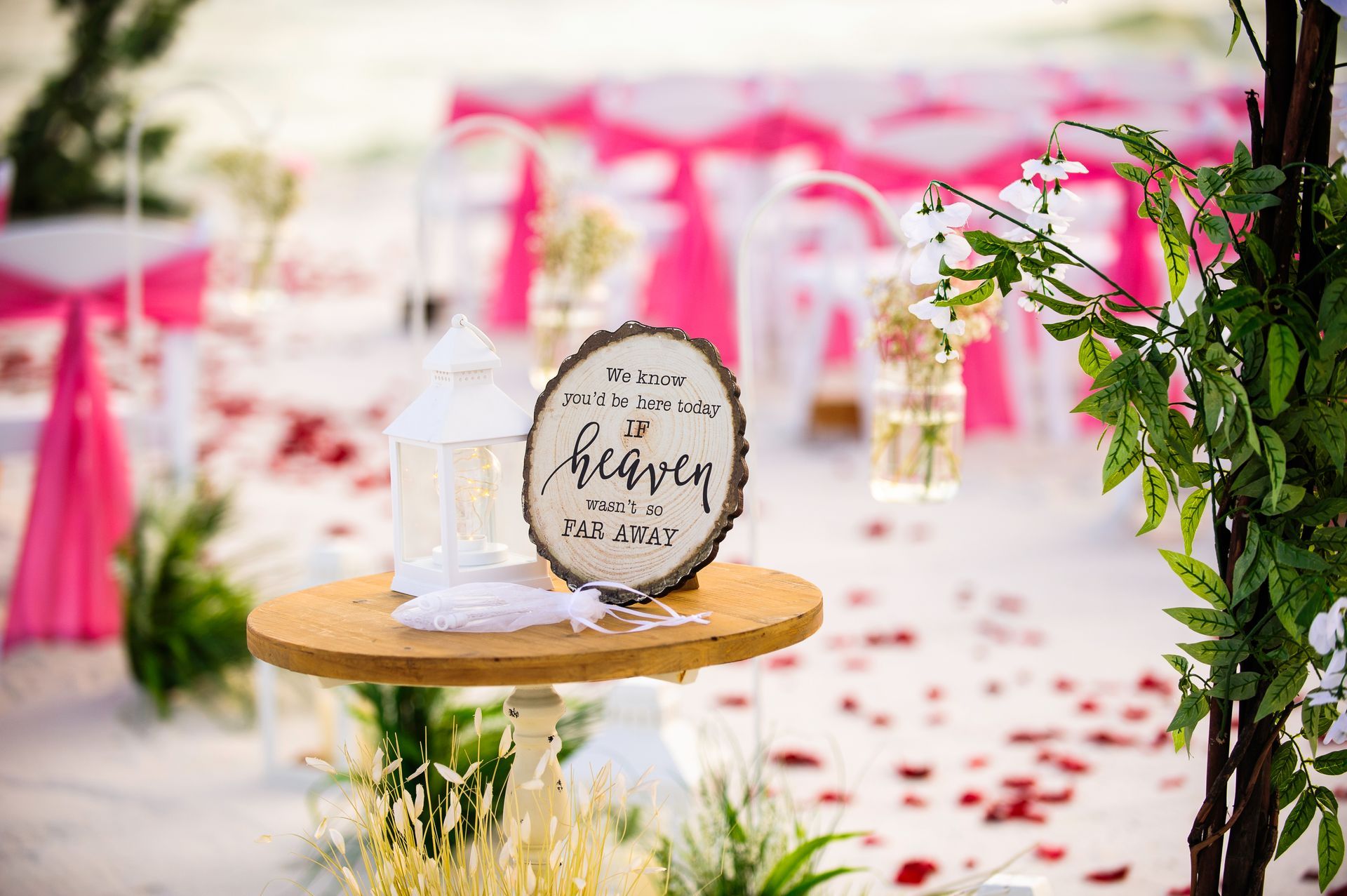 Round wooden sign on a stand in a pink-and-white outdoor wedding aisle with rose petals and flowers