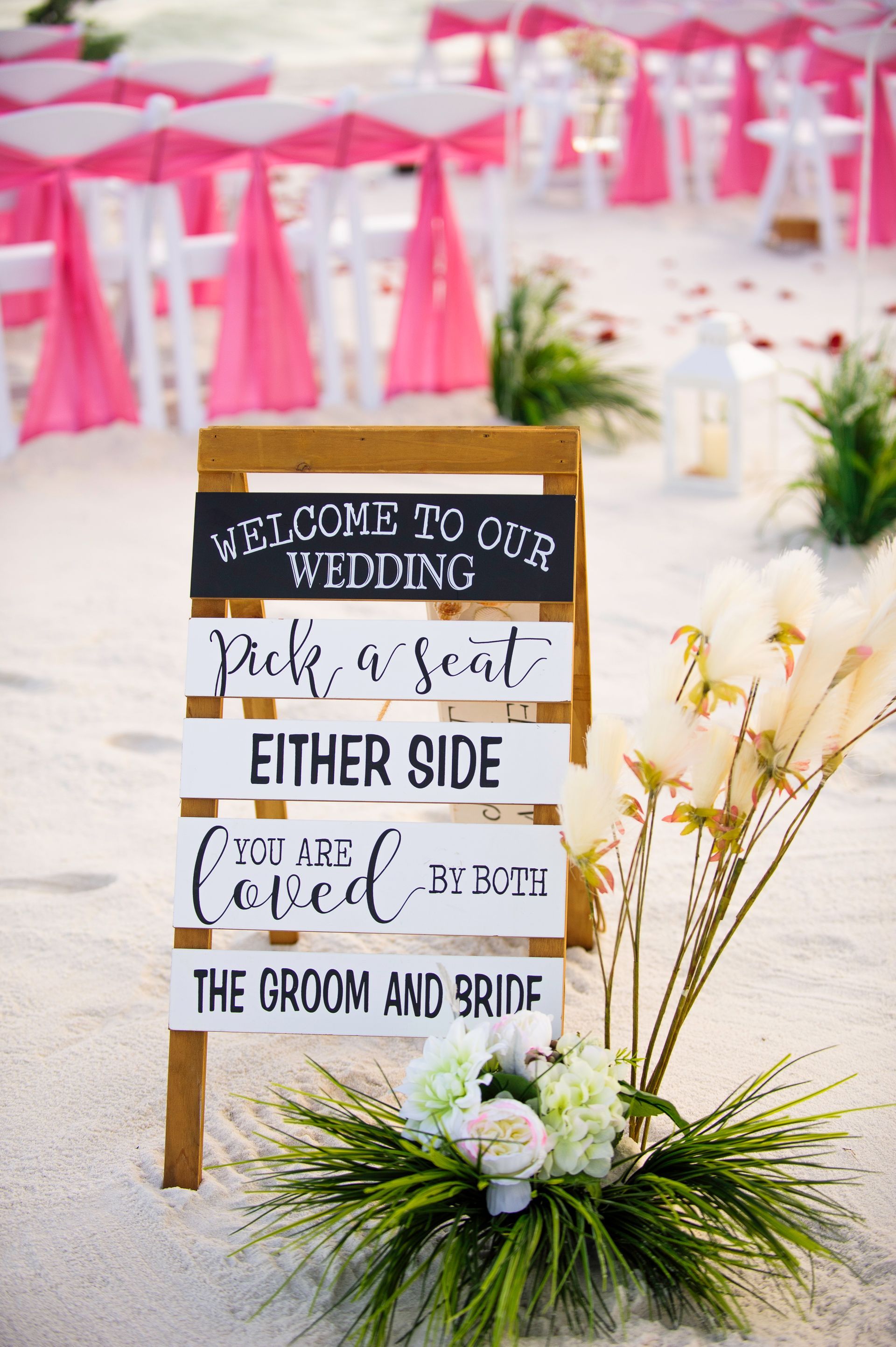 Wedding welcome sign in white and gold on a stand, with pink decor and white chairs in the background