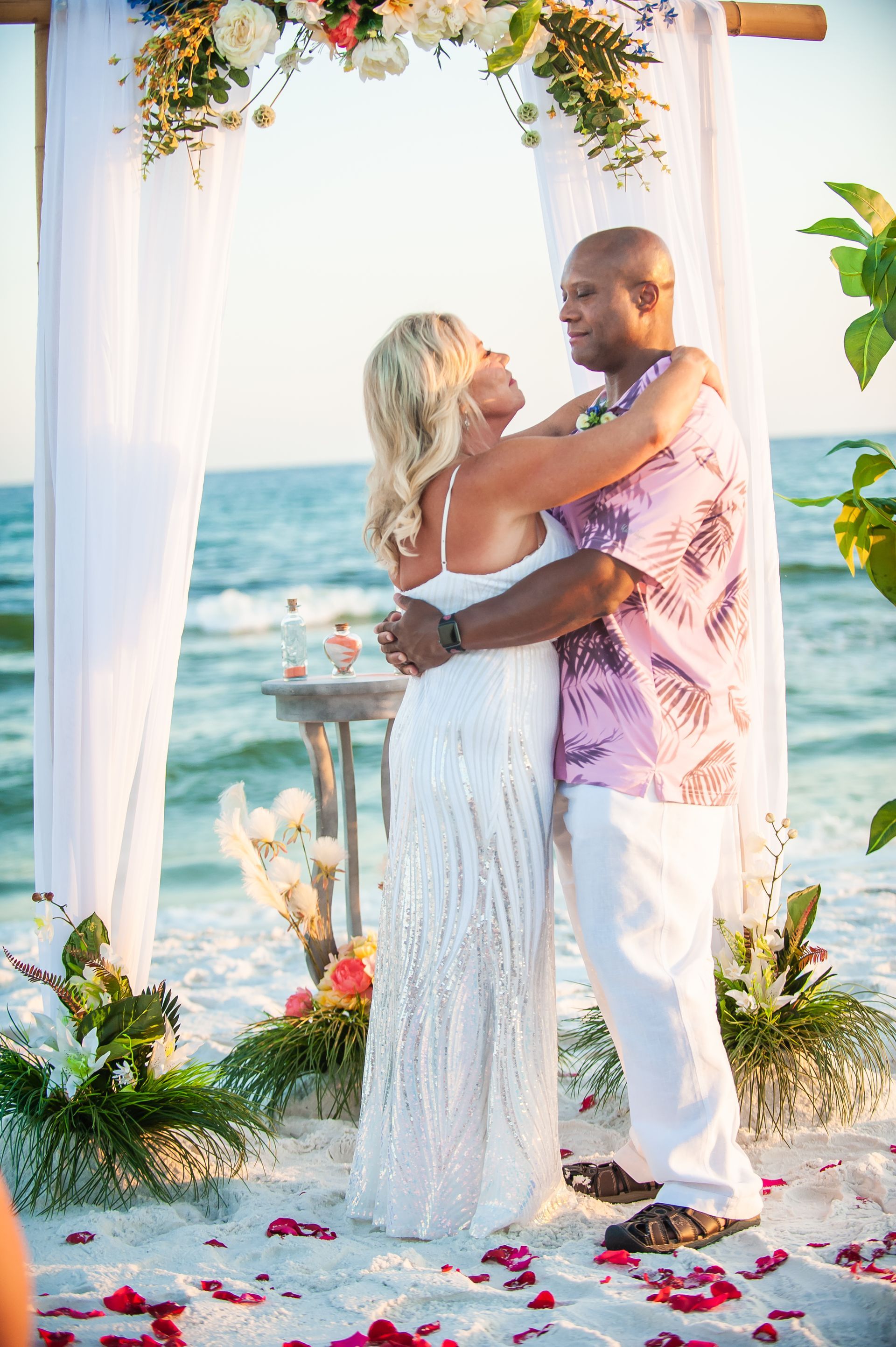 Couple embracing under a floral arch on a beach wedding aisle, with ocean waves in the background.