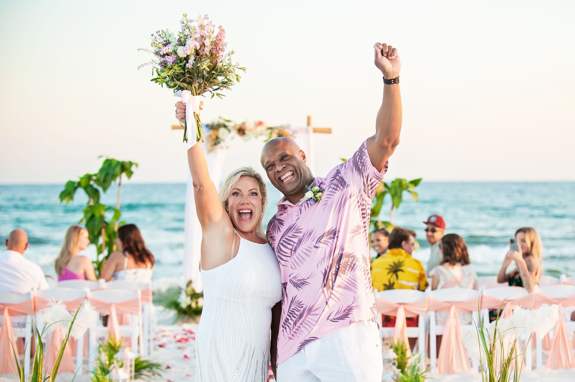 Couple celebrating on a beach at sunset, smiling and holding a bouquet with guests seated behind them