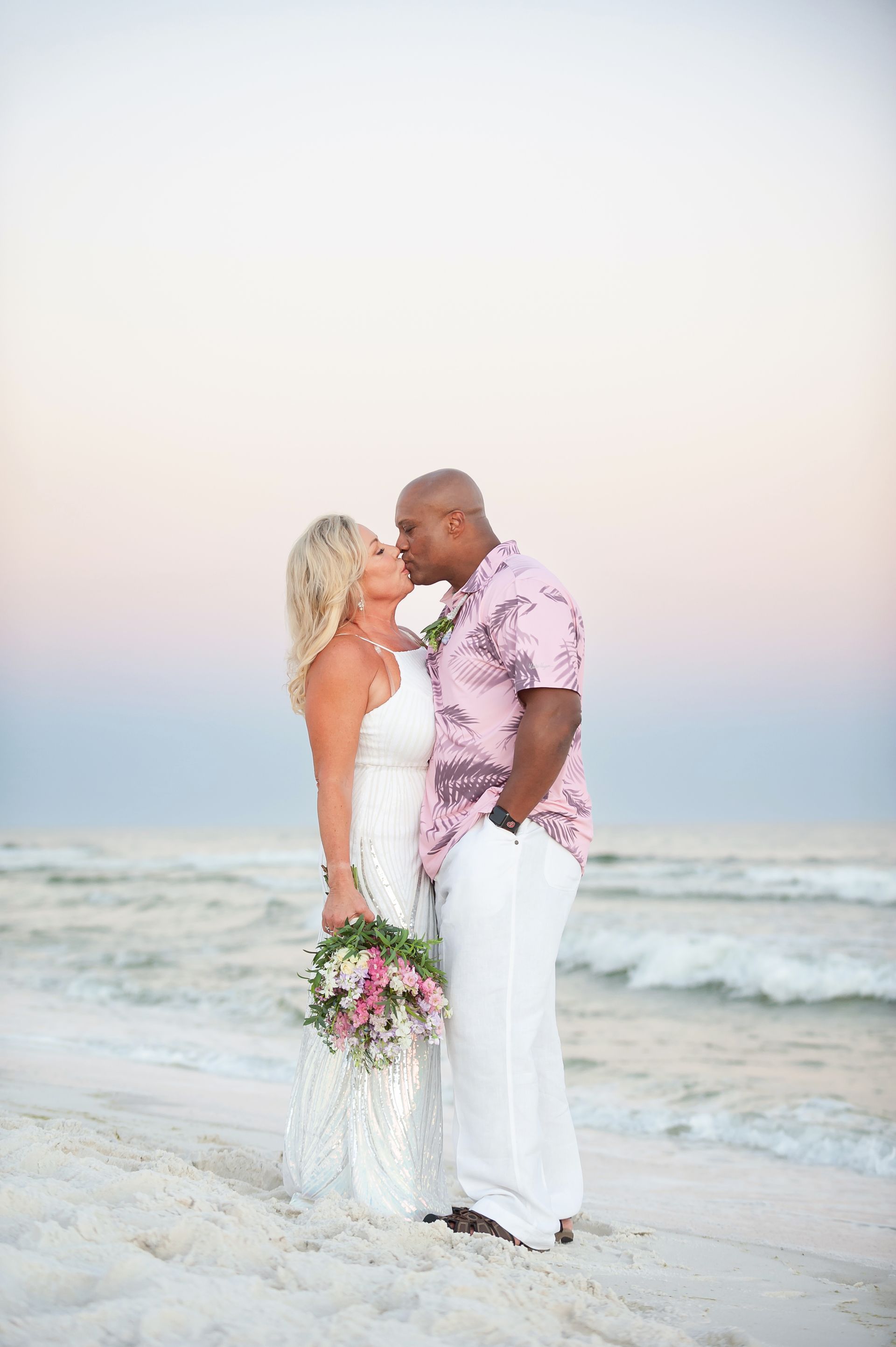Couple in white wedding attire standing barefoot on a beach at sunset, holding each other close.