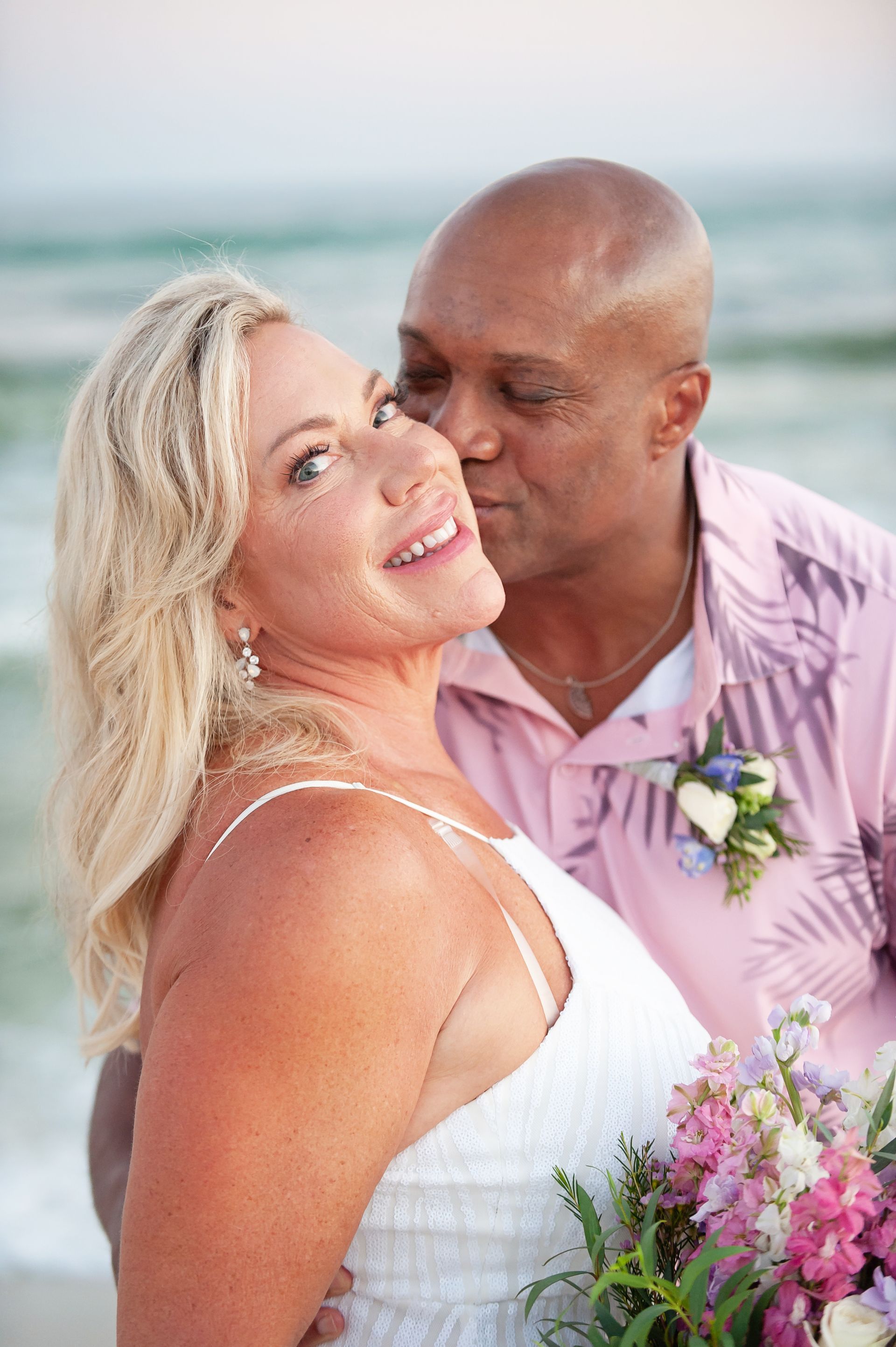 Couple in wedding attire embracing on a beach, with flowers and ocean in the background.