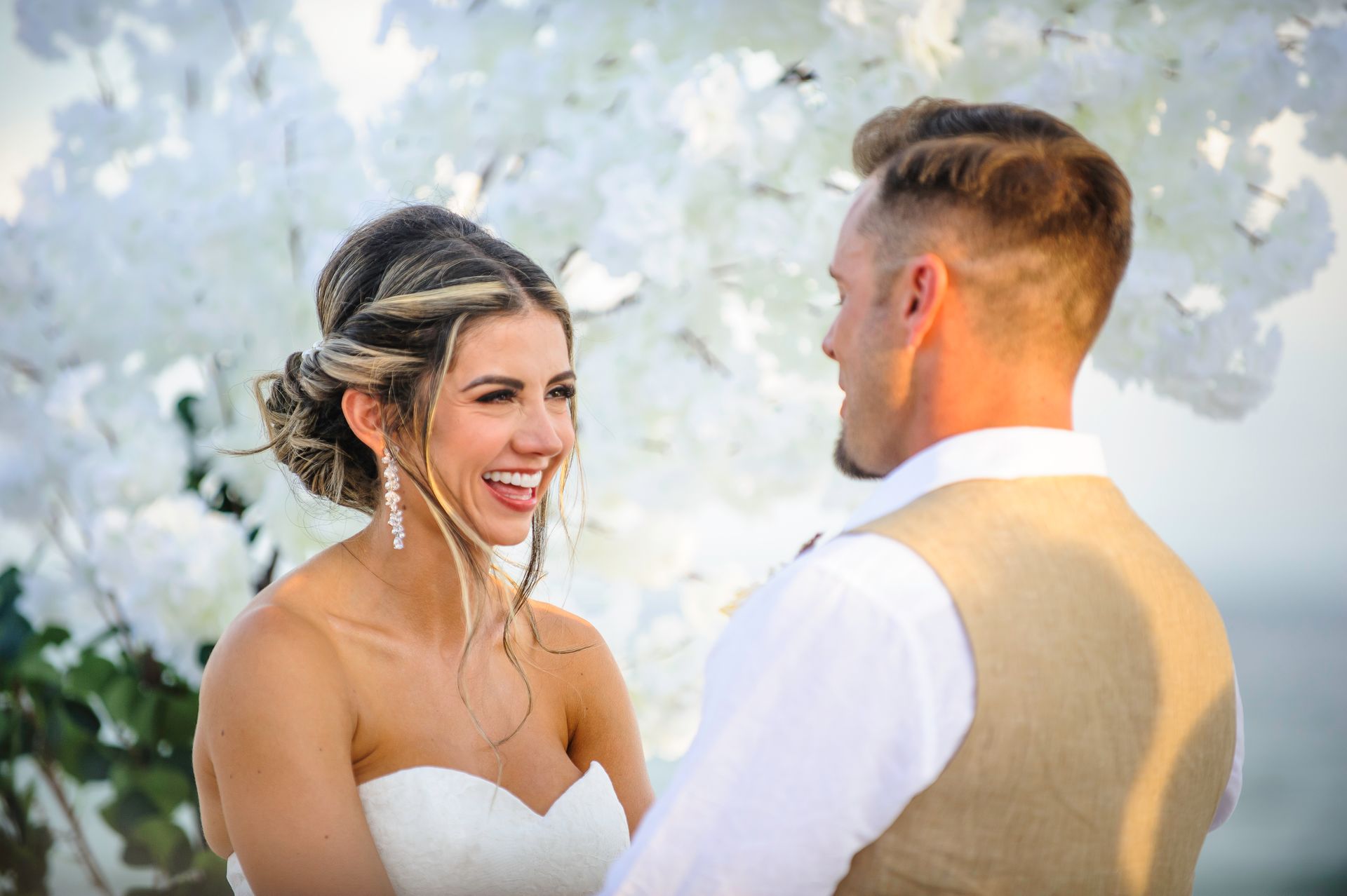 Bride and groom smiling at each other in front of white flowers outdoors