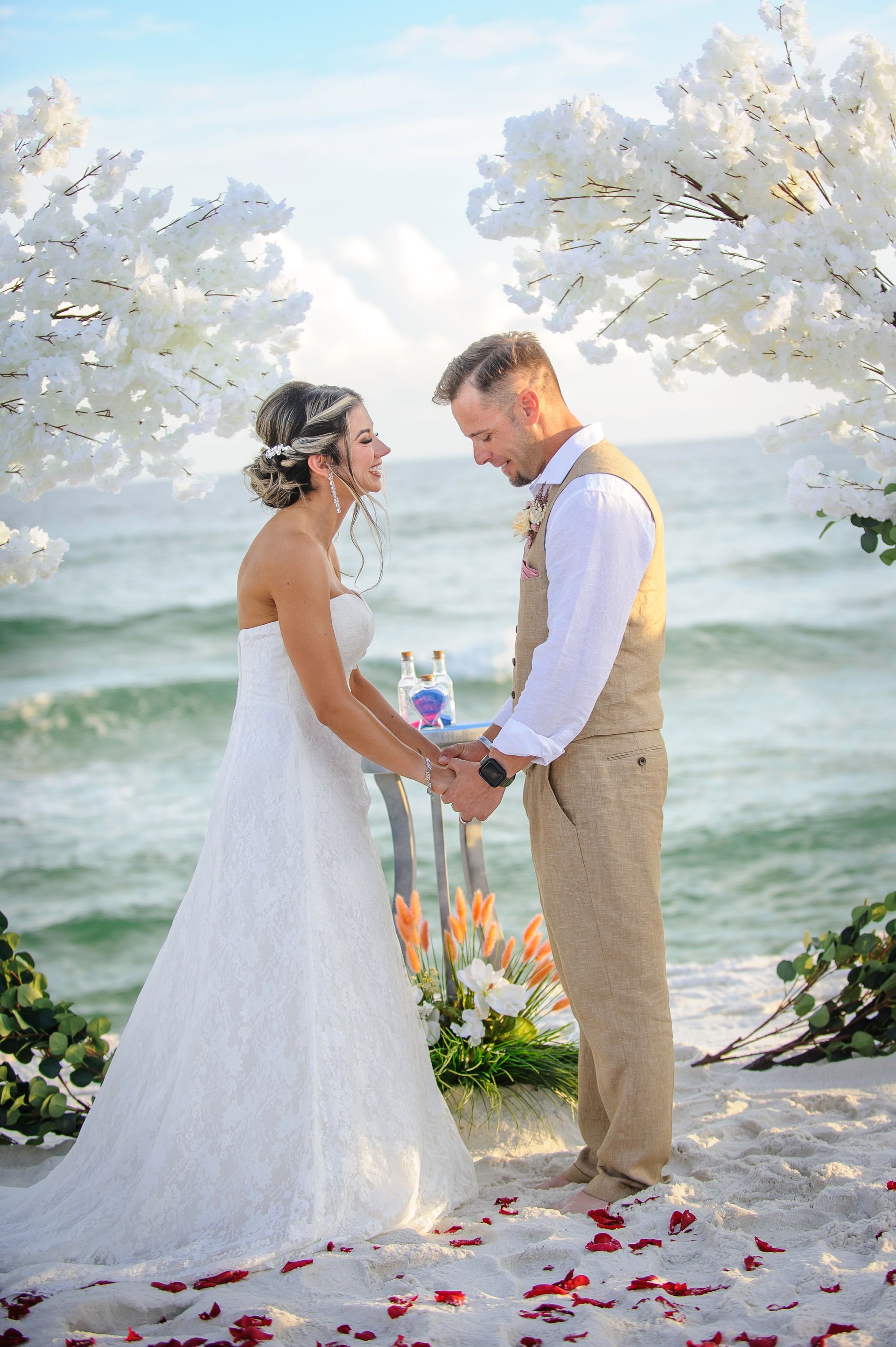 Wedding couple holding hands on a flower-strewn beach, framed by white blossoms and the ocean.