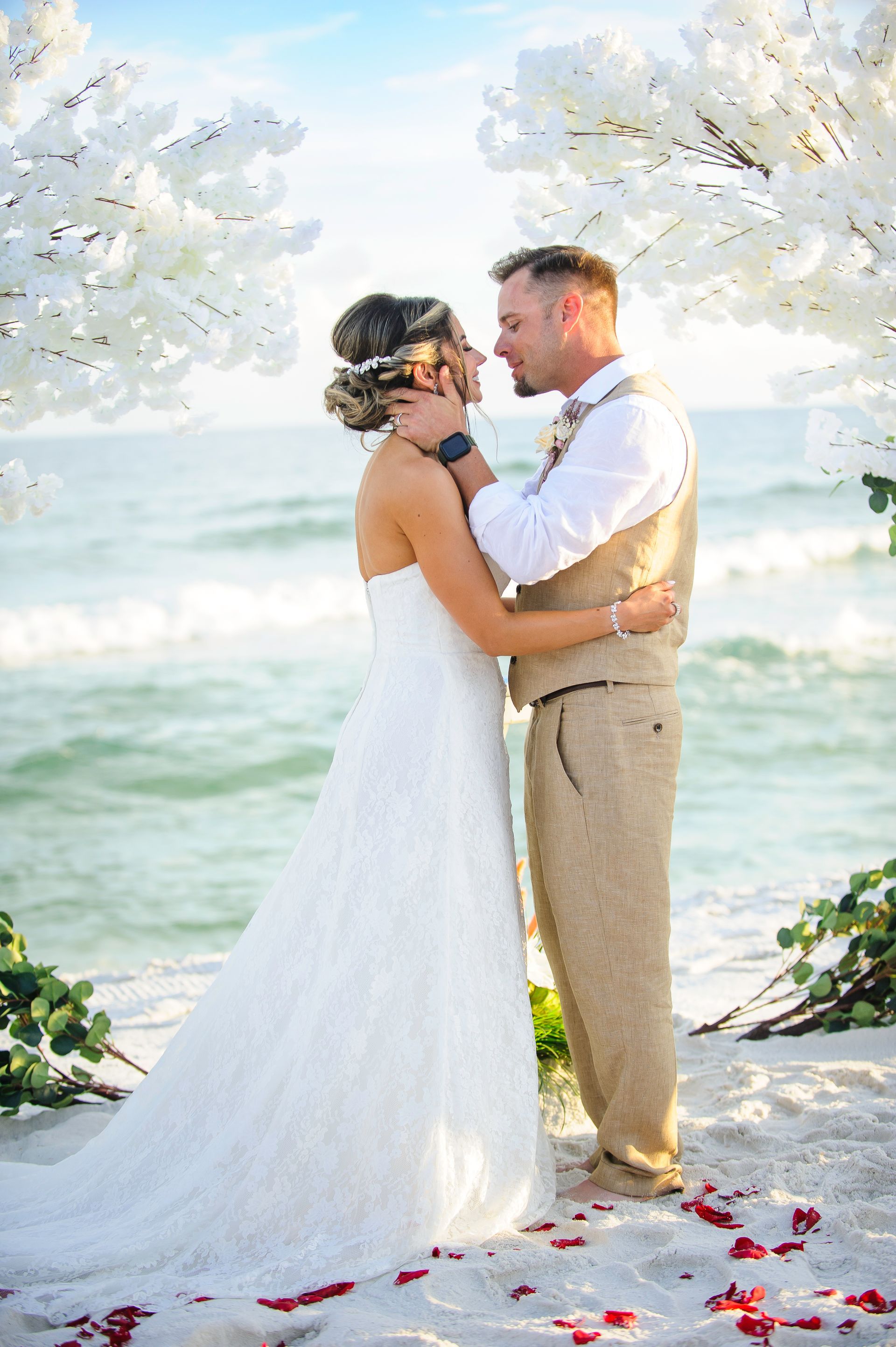 Bride and groom embracing on a beach with white flowers and ocean waves in the background
