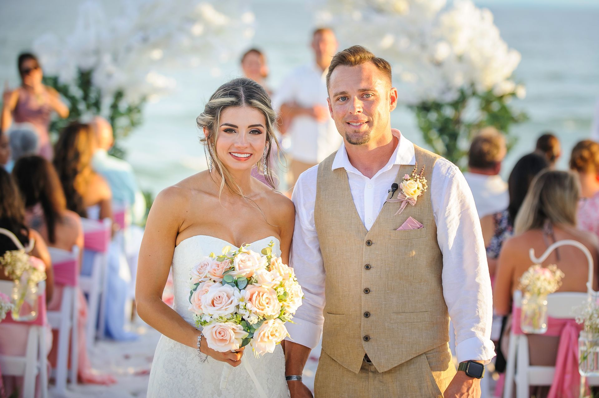 Bride and groom standing at an outdoor wedding, with floral bouquets and guests in the background