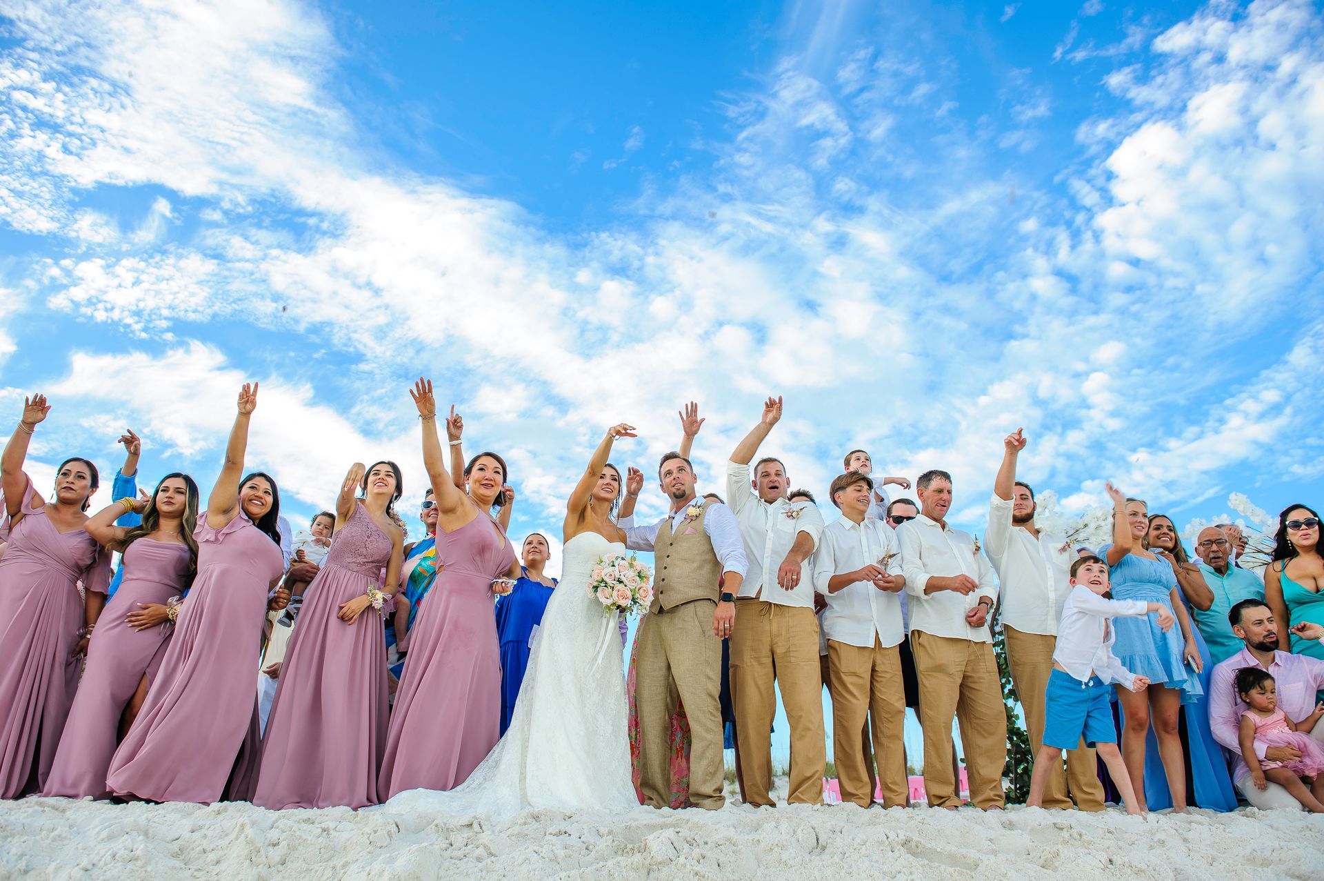 Wedding party posing on a beach, cheering under a bright blue sky