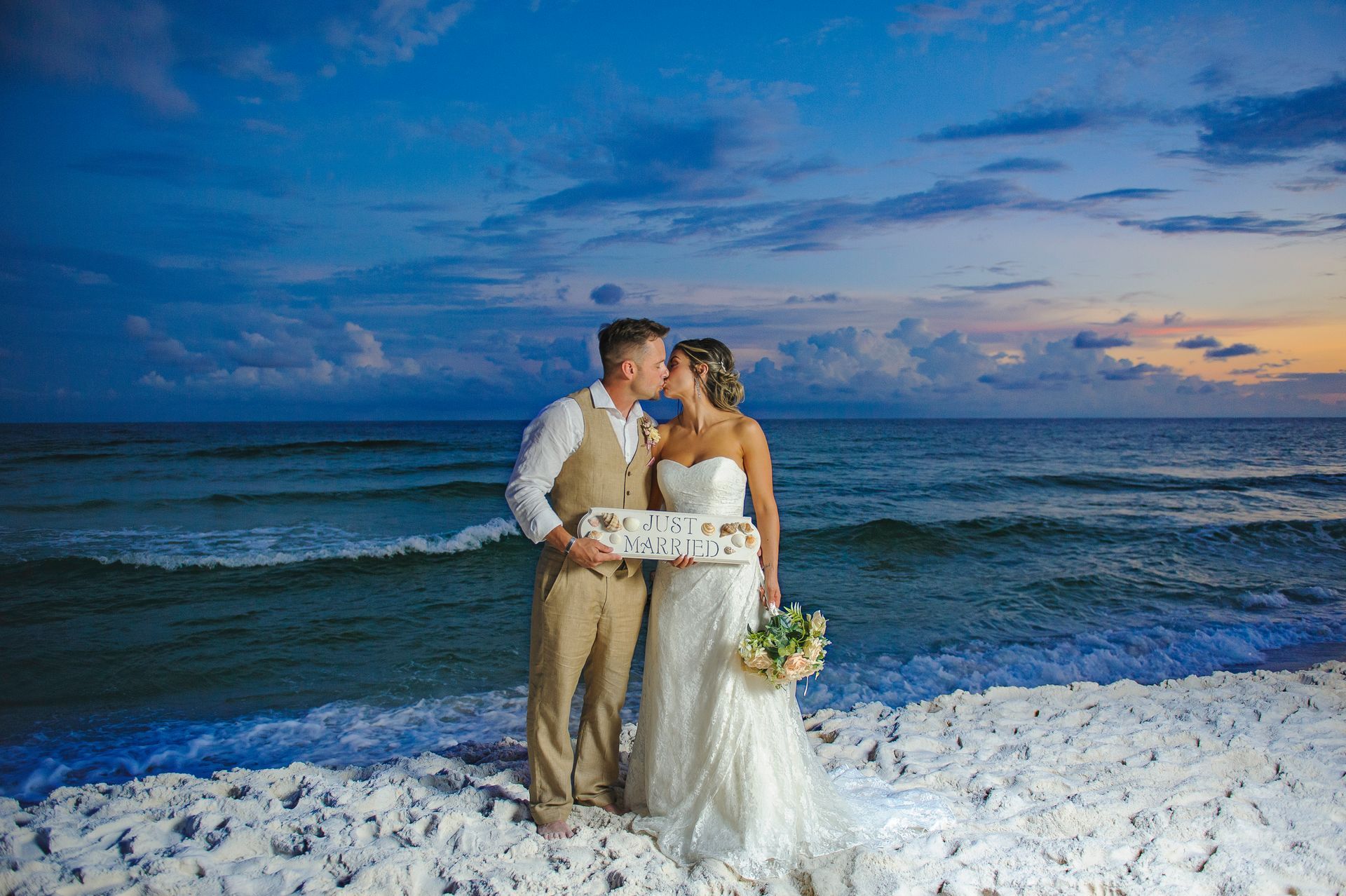 Couple in white wedding attire standing by the ocean at sunset, holding a bouquet on a rocky shore.