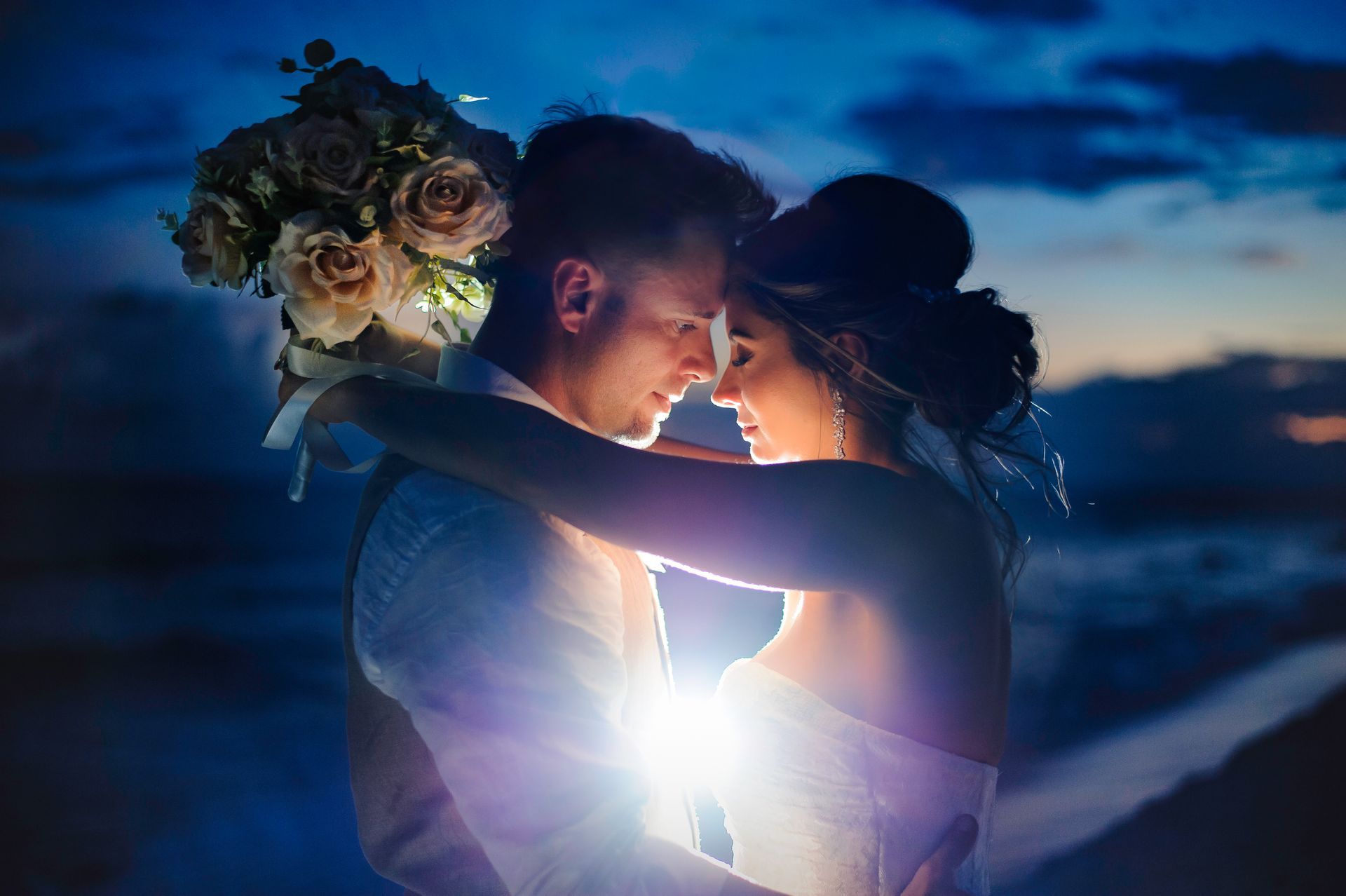 Couple embracing at dusk, backlit by a bright light, with a bouquet and ocean in the background