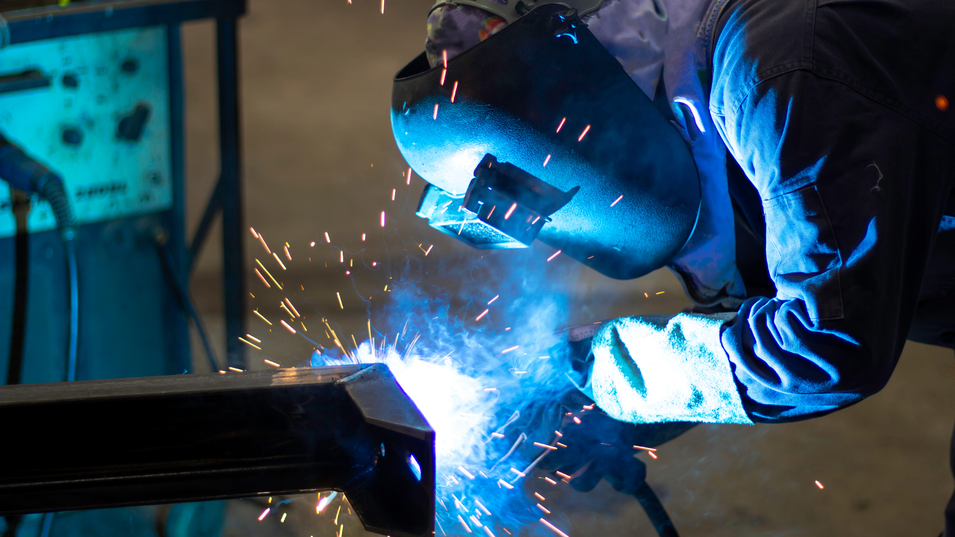 A person in work gloves using a drill press to create a hole in a metal beam, metal shavings visible.