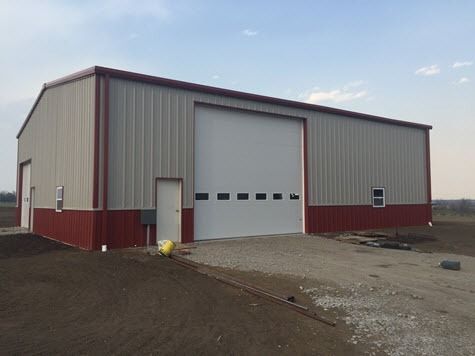 A large, red and tan metal building with a large garage door and small entry door on a gravel driveway.