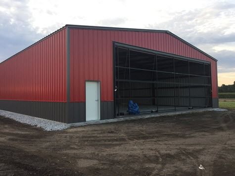 Red and gray metal building with large open doorway, white door, and gravel base.