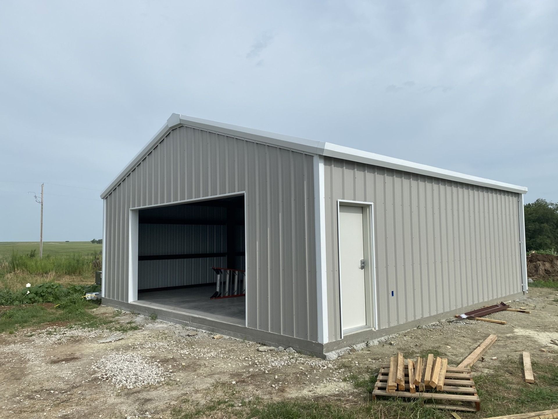 Gray metal building with large garage door and white entry door, under overcast sky.