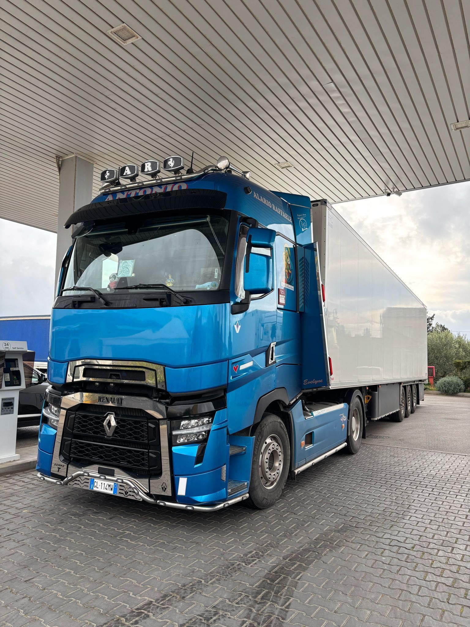 Un camion blu con un rimorchio bianco è parcheggiato in una stazione di servizio.