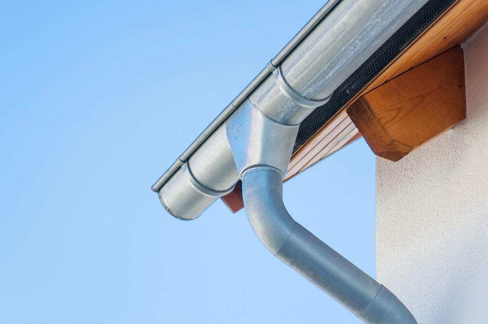 Grondaia e pluviale in metallo grigio su una casa bianca, contro un cielo azzurro.