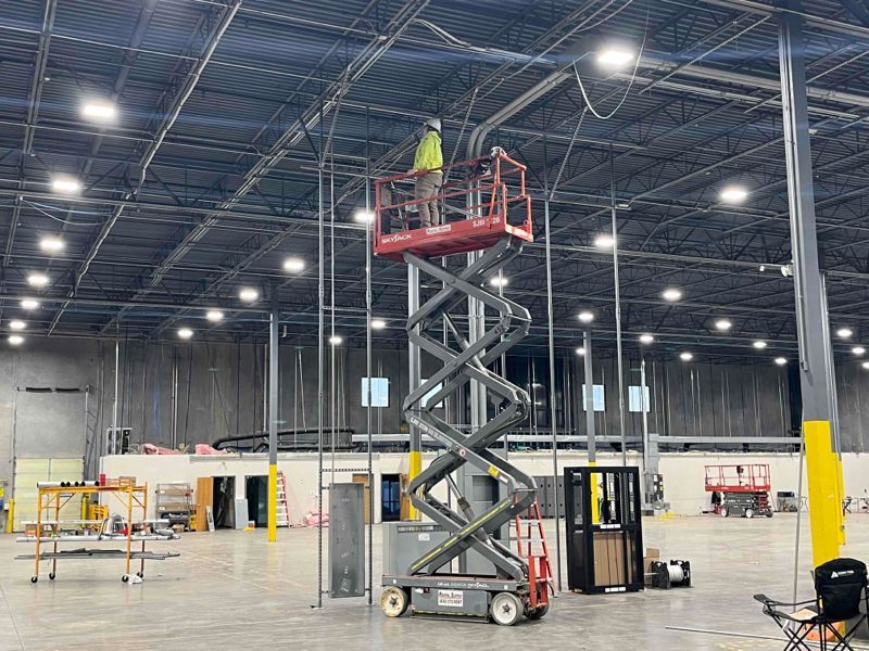 Electrical worker on a raised scissor lift in a large warehouse interior.