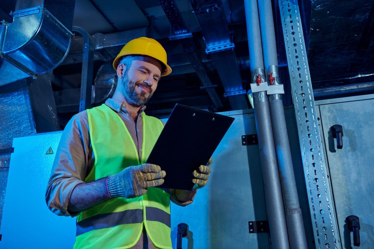 Worker in hard hat and safety vest reviewing a clipboard in a blue-lit industrial facility