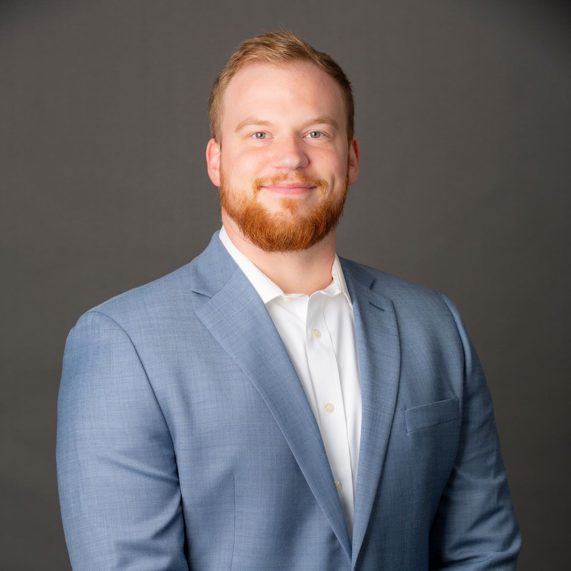 Man with ginger beard wearing a light blue blazer and white shirt, smiling. Gray background.