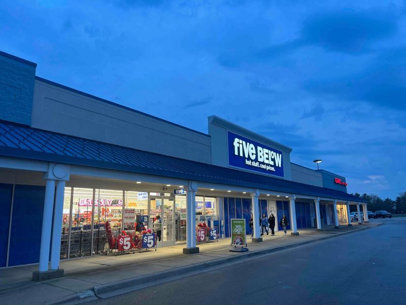 Exterior of a Five Below store with lit windows and shoppers. Blue roof and signage against a dusky sky.