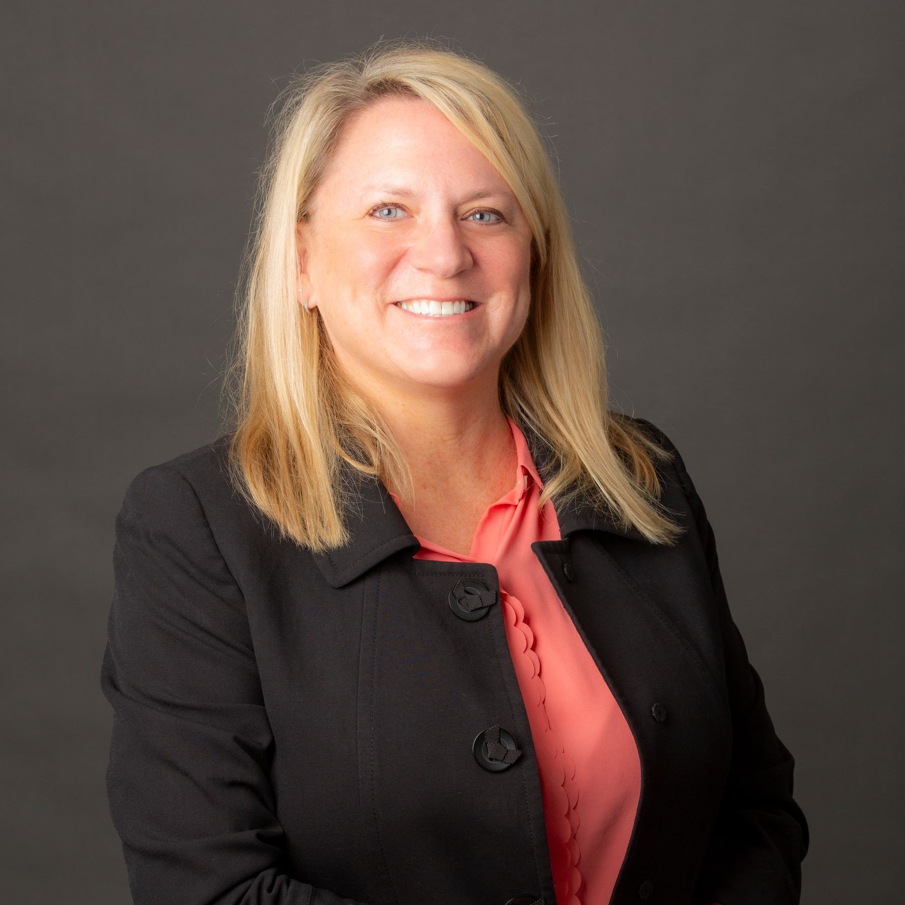 Woman with blonde hair, wearing a black blazer over a coral blouse, smiling, against a gray backdrop.