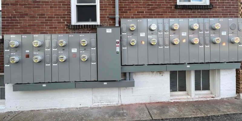 Row of electric utility meters mounted on a brick building wall, with gray panels and basement windows below