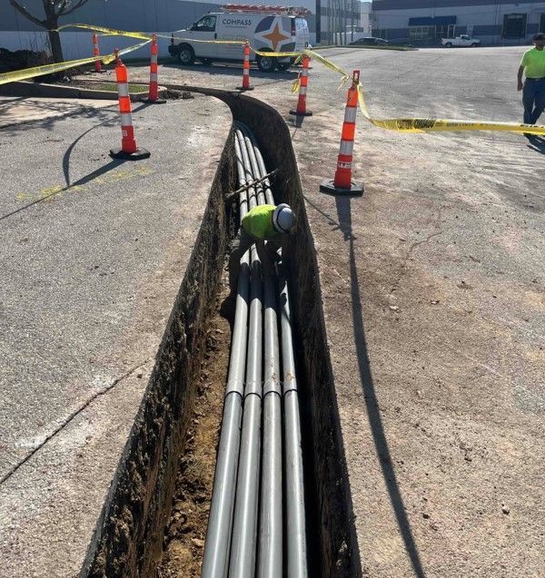 Worker in trench with gray pipes; orange cones and caution tape mark a construction site.
