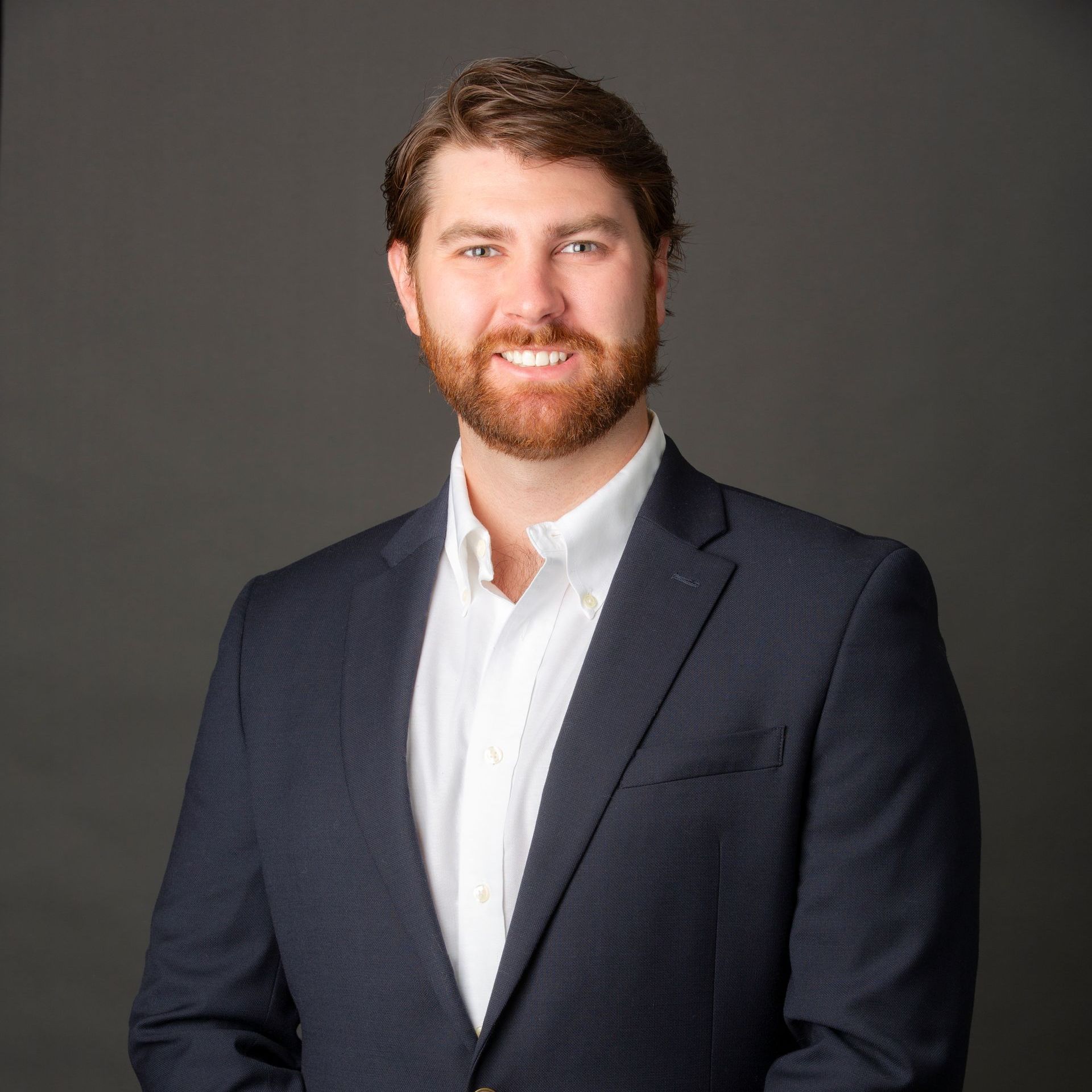 Man with beard in navy blazer and white shirt smiles at the camera. Grey backdrop.