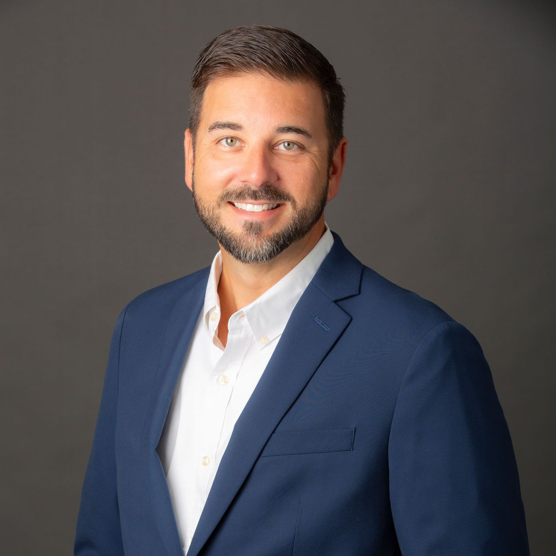 Man in a blue blazer and white shirt, smiling, against a gray background.