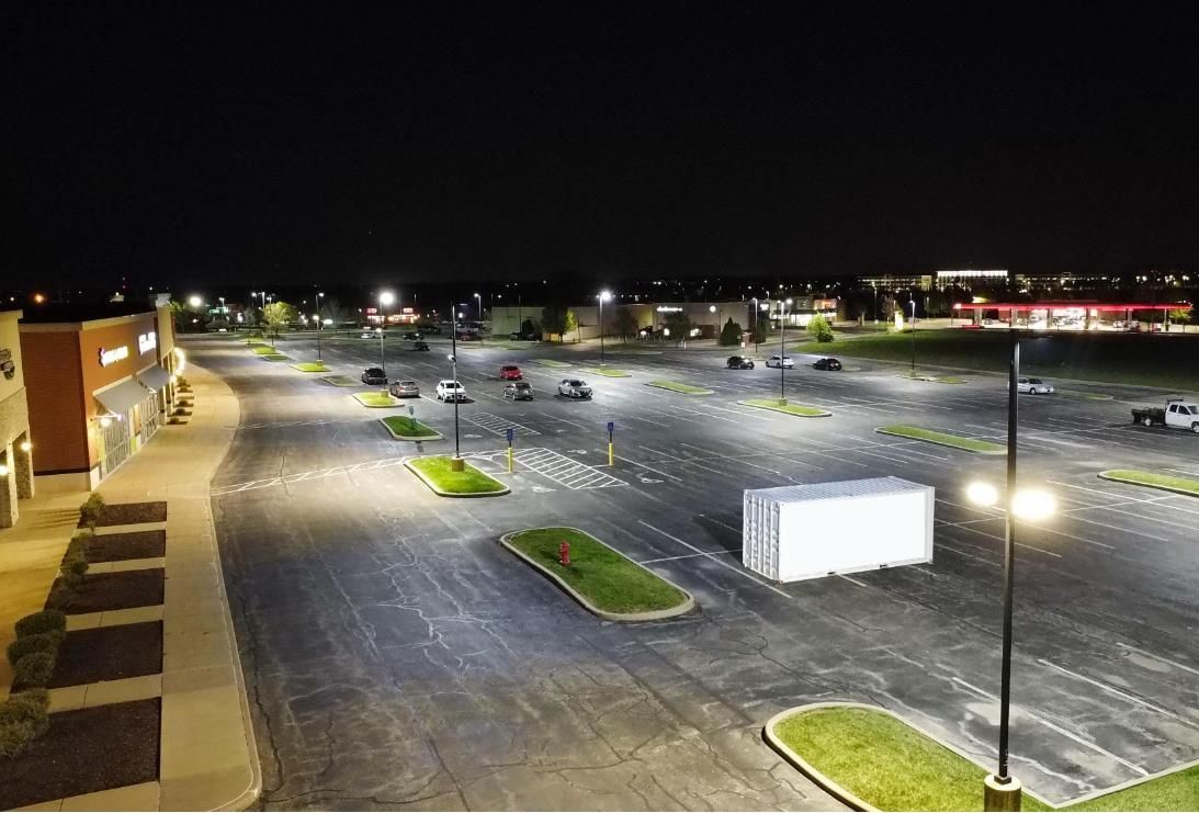 Nighttime aerial view of a mostly empty parking lot illuminated by bright lights. Buildings and a few cars are visible.