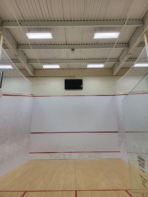 Squash court interior: white walls, wooden floor, red lines, scoreboard, overhead lighting.