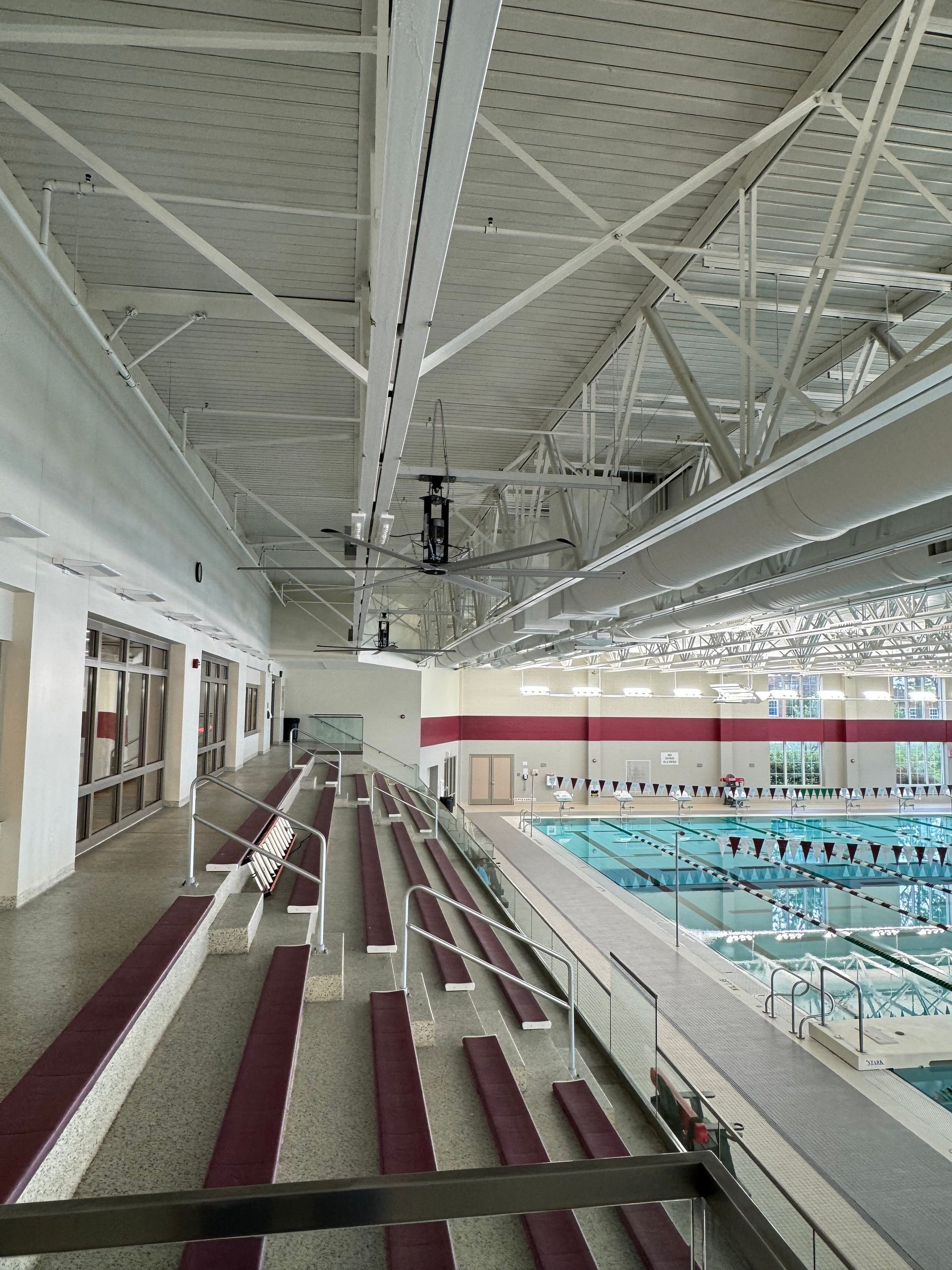 Indoor swimming pool with empty bleachers and a high white ceiling with fans installed; lanes visible below.