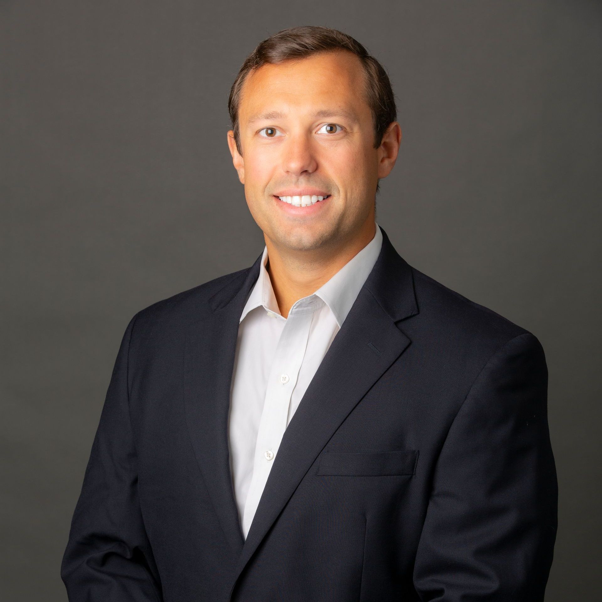 Man in dark suit and white shirt smiles at the camera against a gray background.