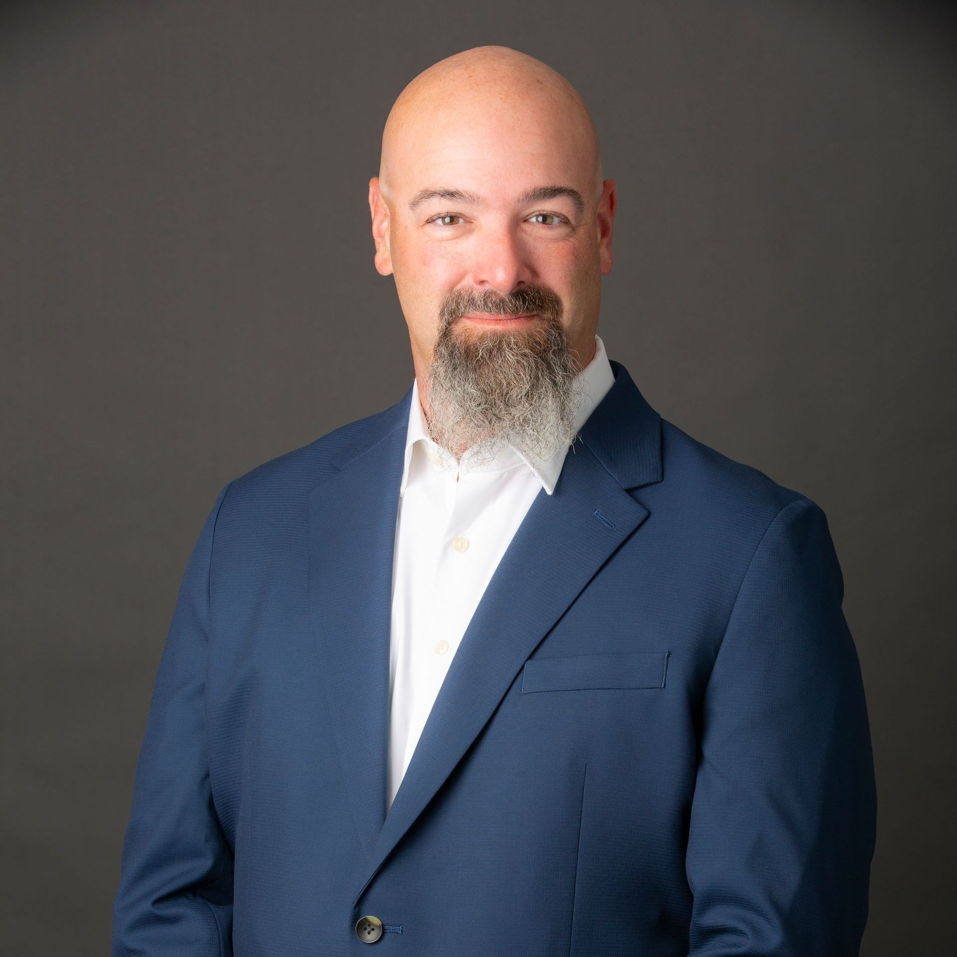 Bald man with a grey beard wearing a blue blazer and white shirt, against a grey background.
