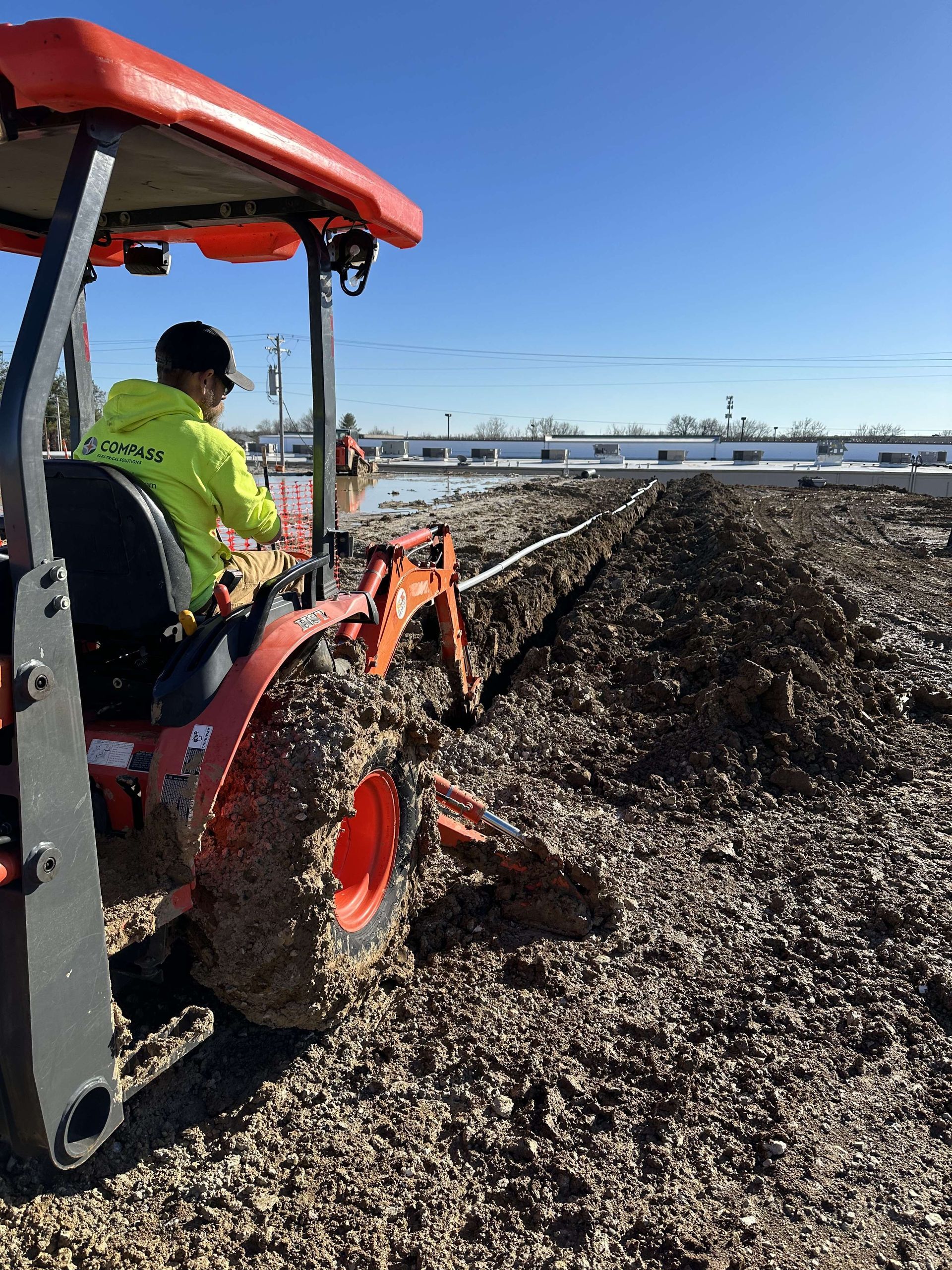 Tractor with backhoe digging in muddy ground; worker in neon green shirt. Sunny day.