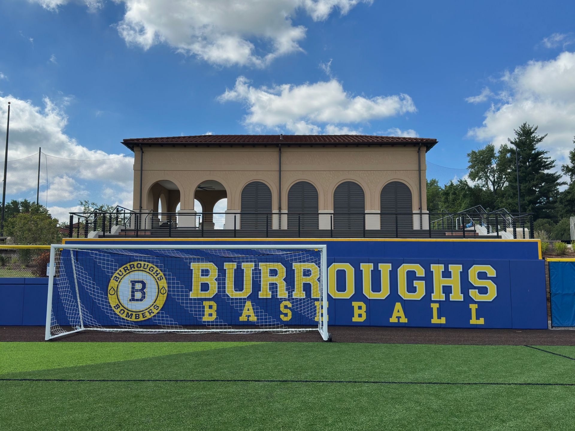 Baseball field with blue and gold "BURROUGHS BASEBALL" sign. Covered structure in the background. Cloudy sky.