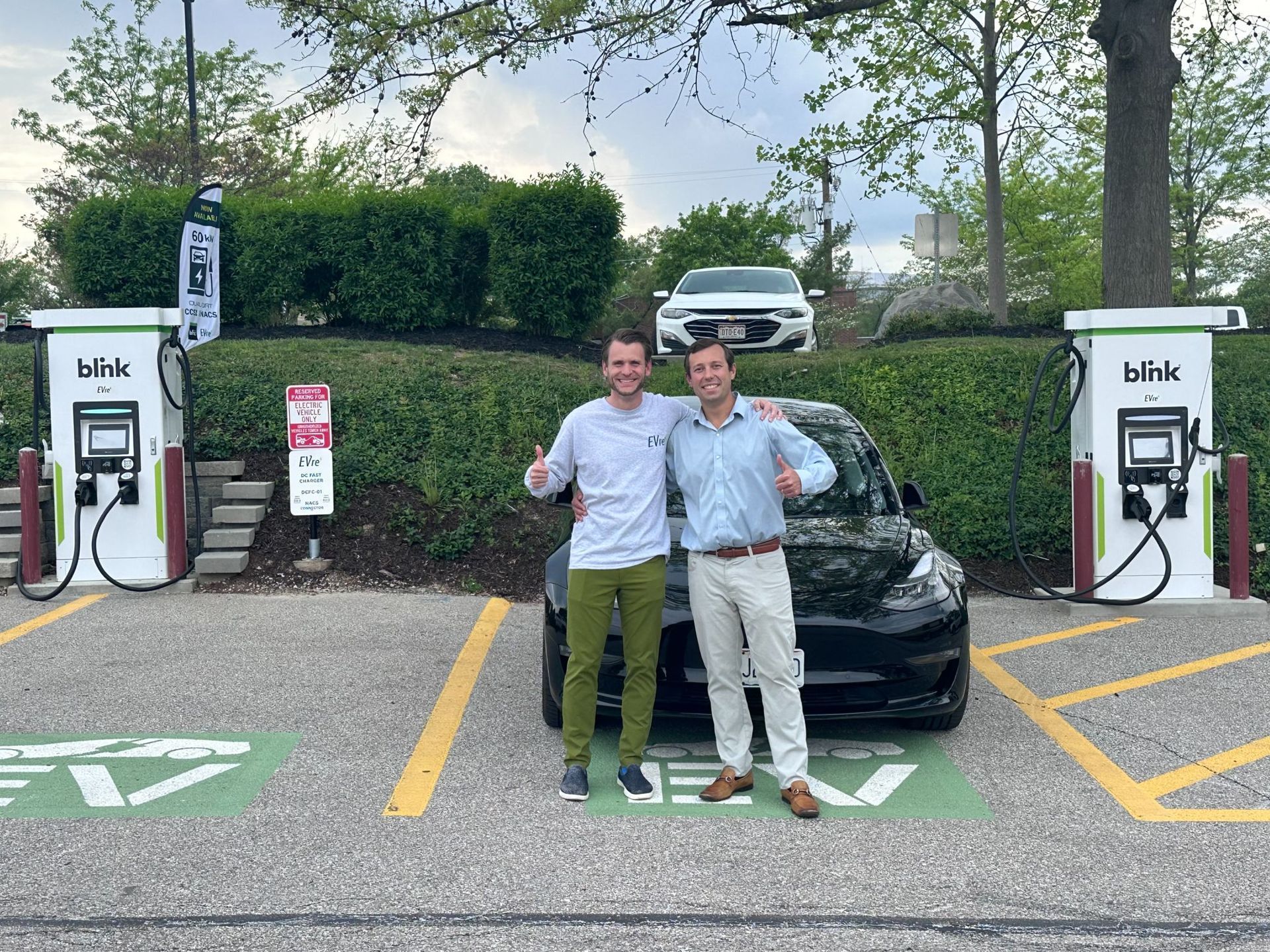 Two men smiling, giving thumbs up next to a black EV charging at a Blink station. Trees and a white car in background.