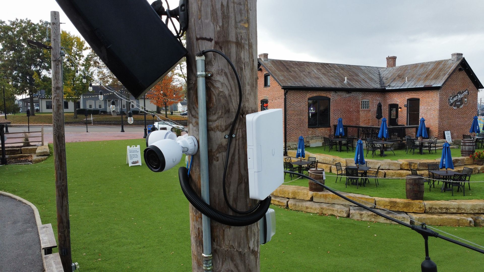 Security camera, speaker, and Wi-Fi access point mounted on a wooden pole near an outdoor seating area with a building backdrop.