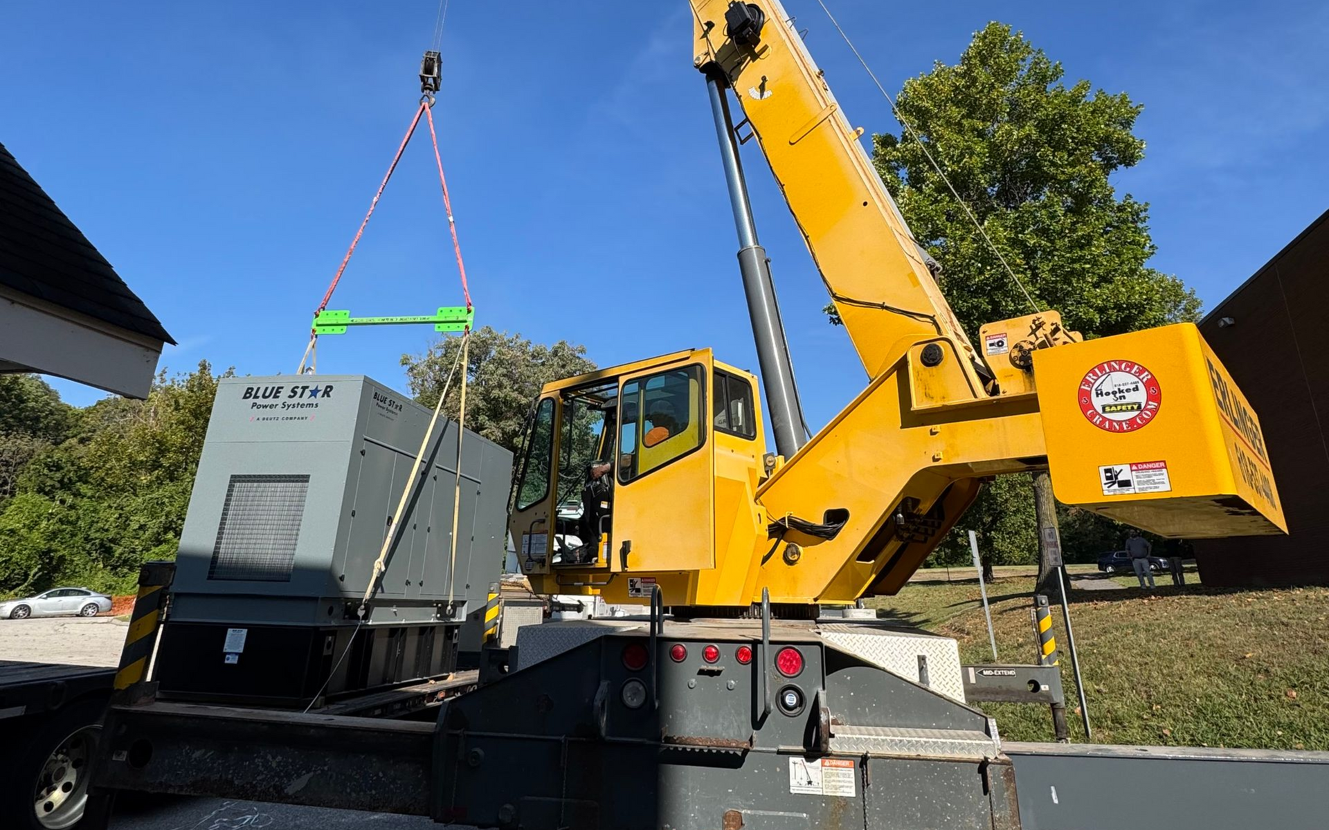 Yellow crane lifting a gray industrial back-up generator unit onto a truck in a sunny outdoor lot