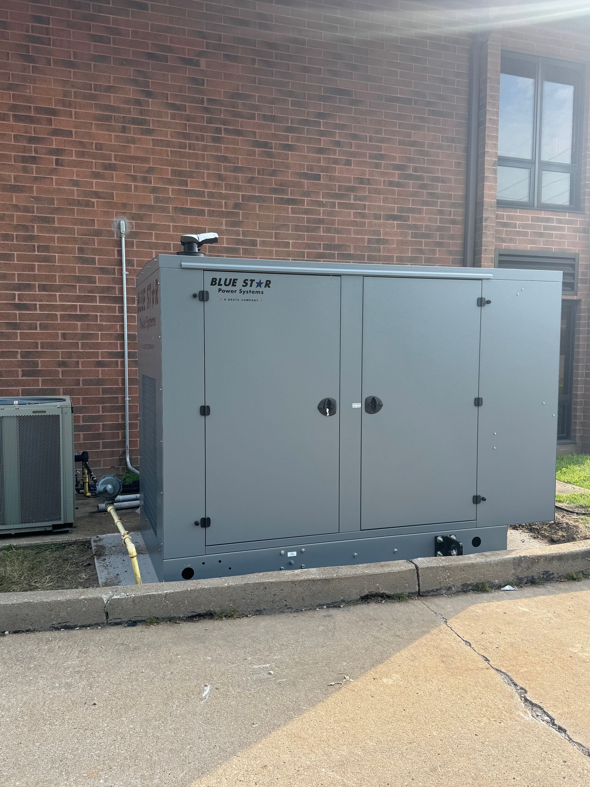 Gray utility cabinet beside a brick building and window, with an AC unit nearby on a paved lot