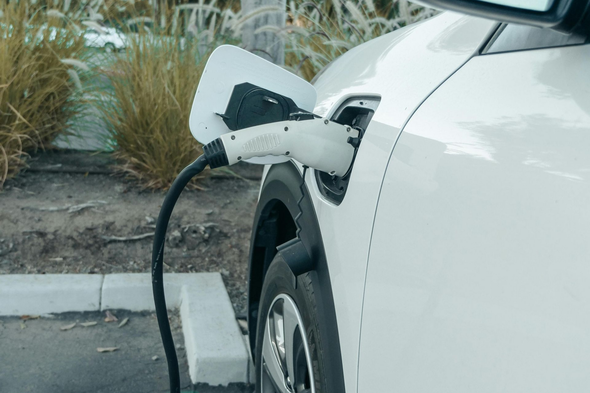 White electric car plugged into a charging station in a parking lot, close-up of the front wheel and cable.