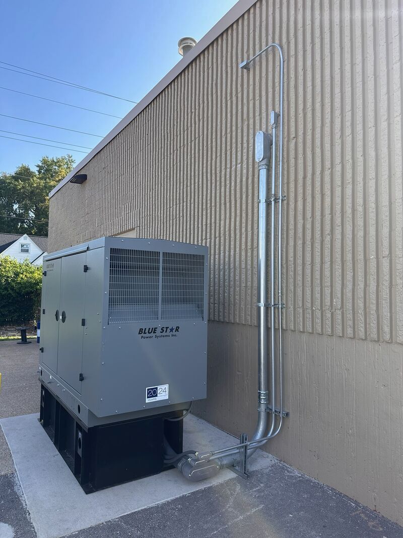 Gray generator next to a building with silver conduit. Concrete pad, blue sky.