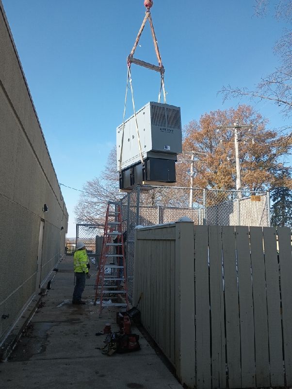 Crane lifting a large back-up generator unit over a fenced construction site with a worker nearby