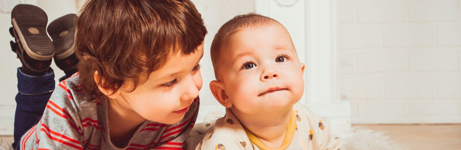 Two children , a boy and a baby , are laying on the floor looking at each other.