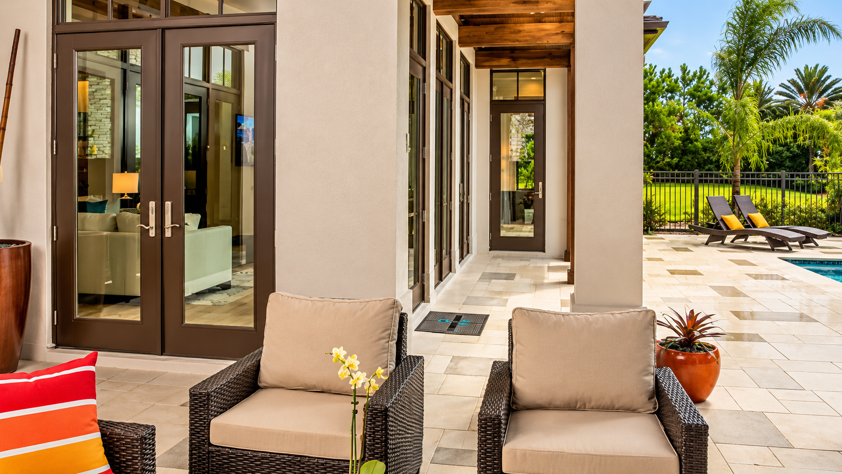 Patio seating beside a villa corridor with glass doors and a view of palm trees and lounge chairs
