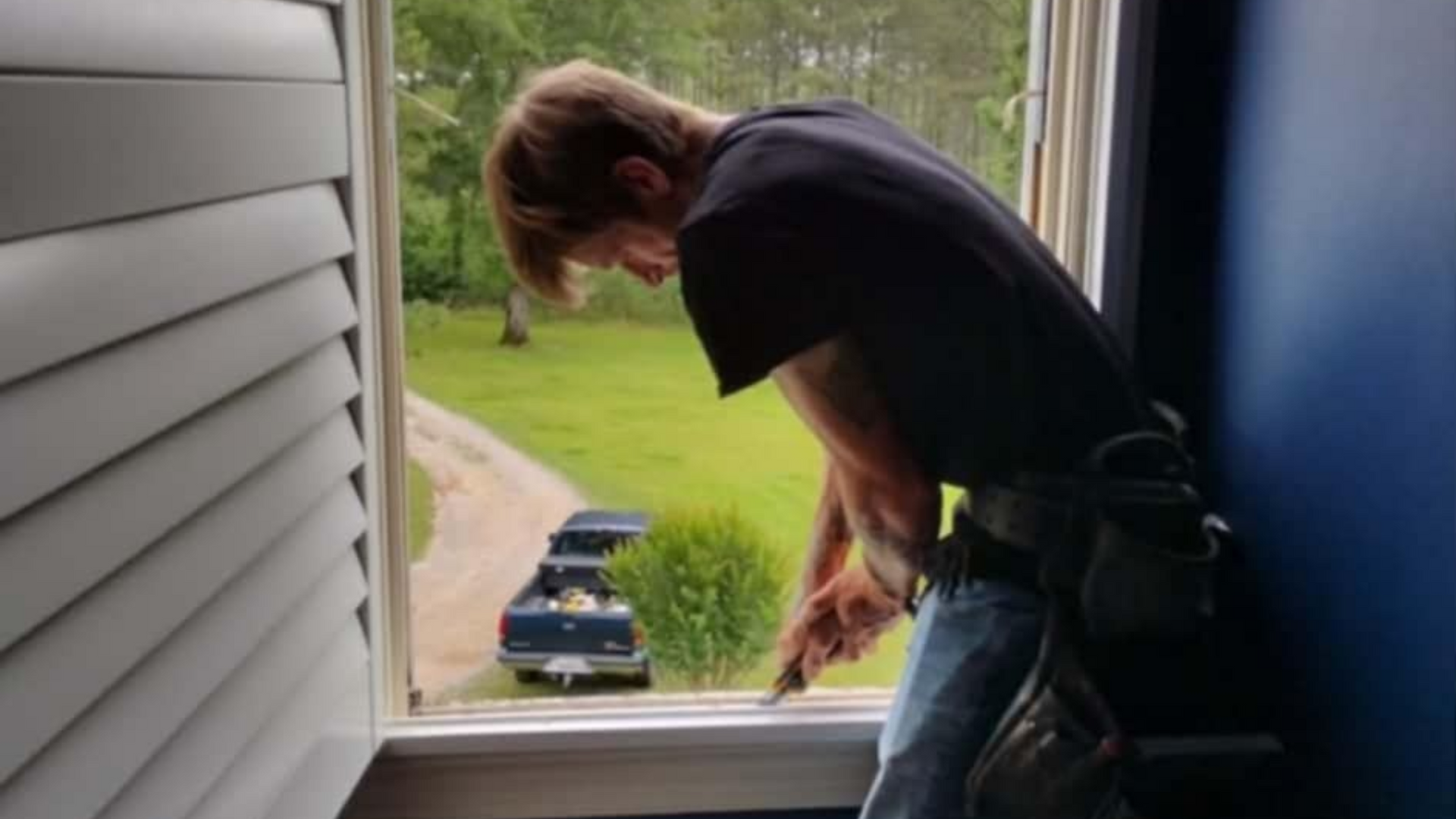 Person leaning out of an open window, working on the sill with a view of trees outside.