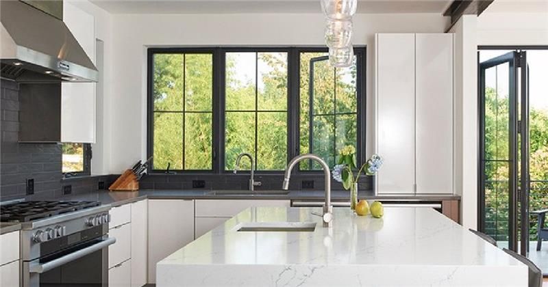 Modern kitchen interior with large black-framed windows, white cabinets, quartz island, and natural outdoor view.